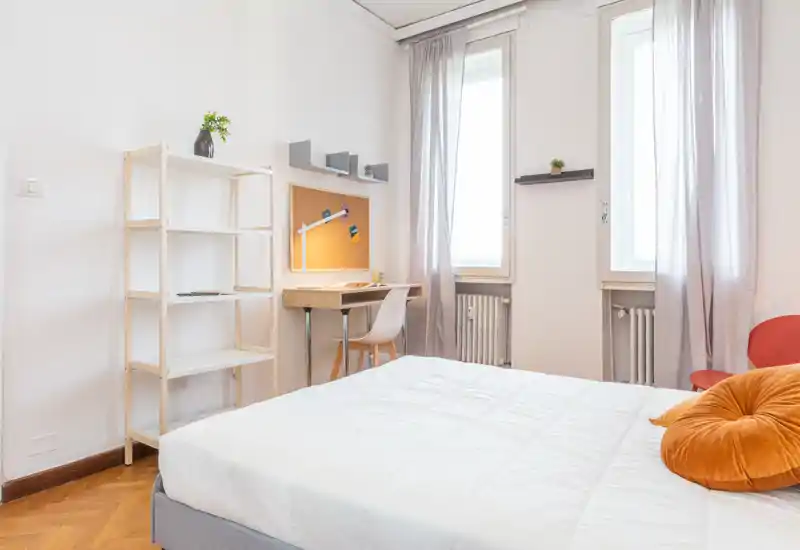 Bright, modern bedroom with a large bed in the foreground, a small desk by the window and soft natural light. Clean minimalist styling with warm accents and practical shelving.