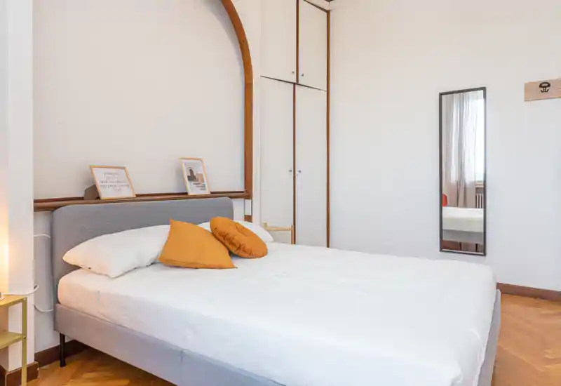 Bright, modern bedroom with a full bed dressed in white linens and warm mustard accent pillows; simple, elegant decor and a full-length mirror on the wall.