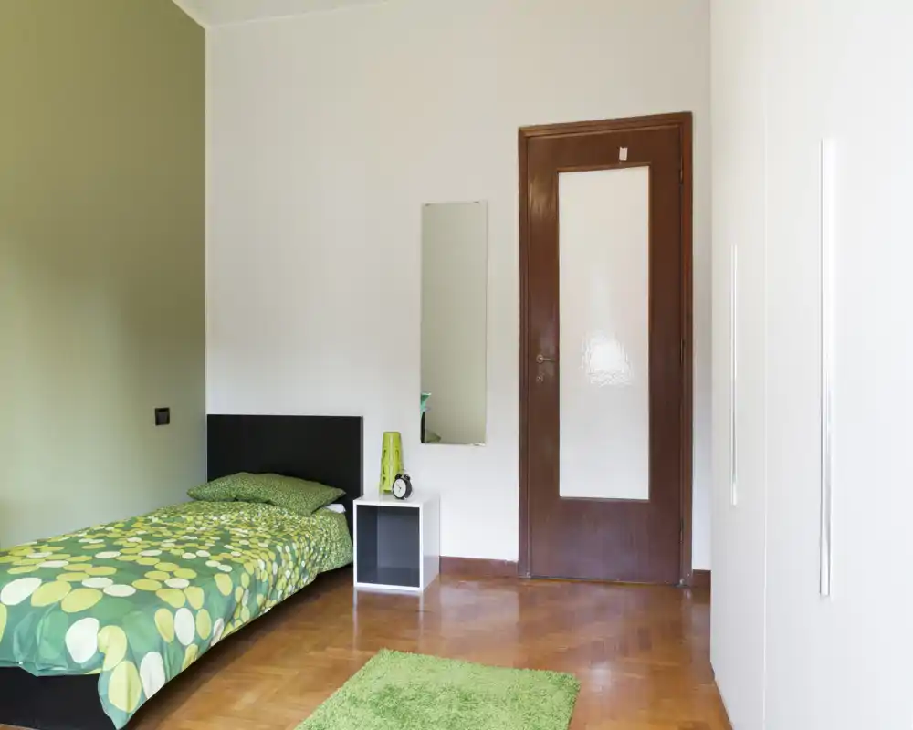 Dormitorio individual luminoso con edredón verde estampado, mesita y espejo de cuerpo entero — ambiente limpio, sencillo y bien iluminado.