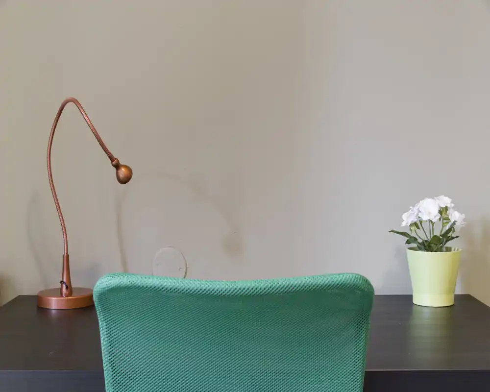 Sencilla zona de trabajo con mesa de madera oscura, silla verde de malla, lámpara de escritorio cobre y una pequeña maceta con flores blancas frente a una pared neutra.