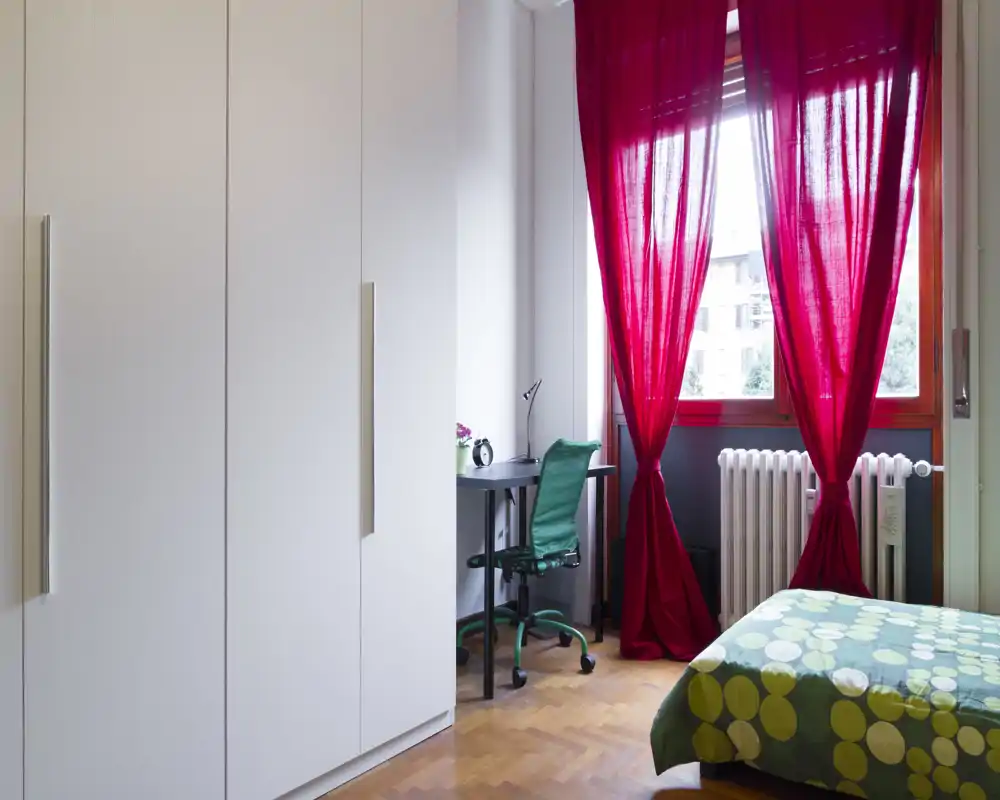 Dormitorio moderno y luminoso con colcha verde estampada, amplio armario blanco y un pequeño escritorio junto a la ventana con cortinas rojas que dejan entrar luz natural.