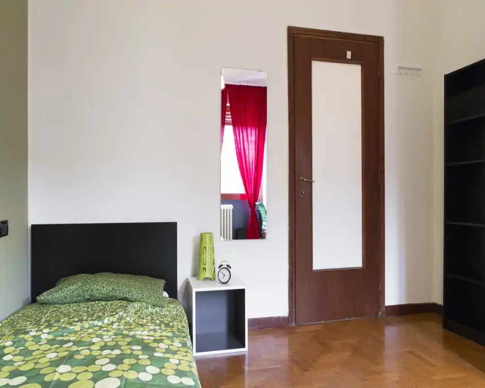 Habitación pequeña y ordenada con cama individual, mesita y estantería alta. Espacio luminoso y sencillo con suelo de parquet y un espejo que refleja una ventana con cortinas rojas.