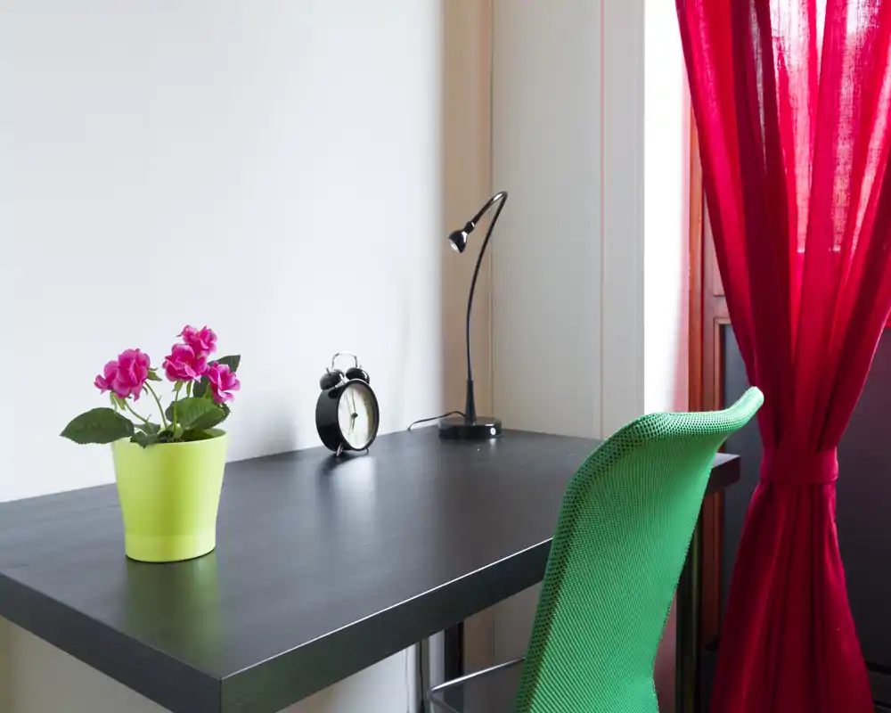 Un compacto y ordenado espacio de trabajo con escritorio negro, lámpara, despertador y una planta rosa junto a una silla verde y una cortina roja.