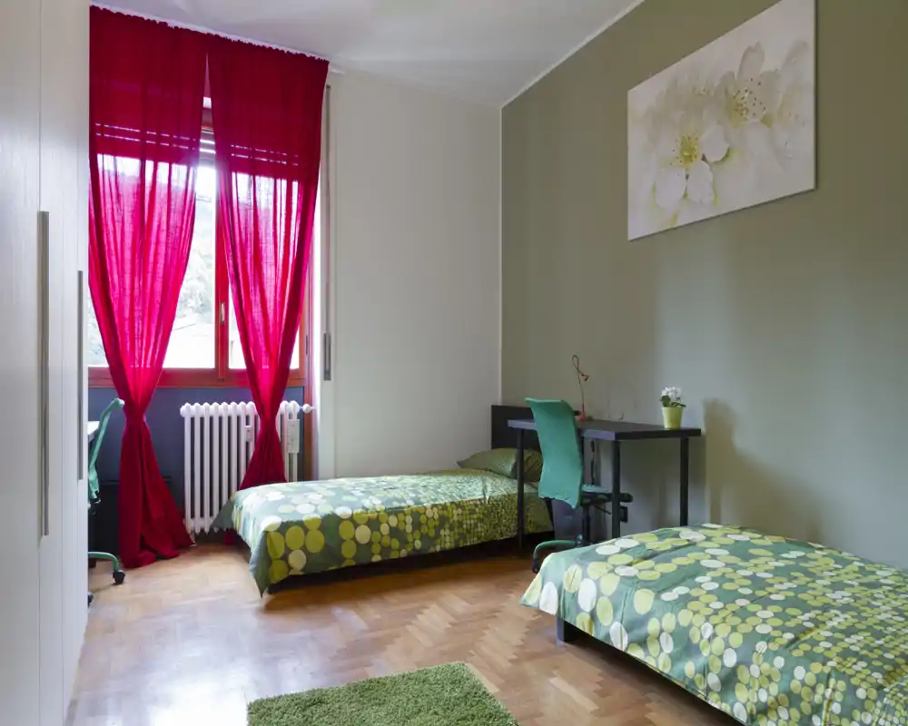 Dormitorio luminoso con dos camas individuales, ropa de cama verde a juego, un escritorio con silla junto a la pared y una gran ventana con cortinas rojas que deja entrar luz natural.