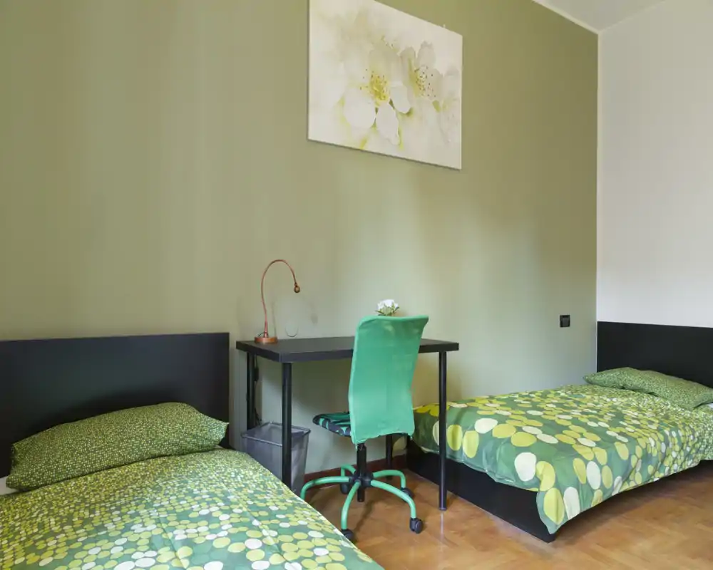 Dormitorio con dos camas individuales, ropa de cama verde a juego y un pequeño escritorio con silla verde. Habitacion ordenada y funcional, perfecta para estancias compartidas o estudiantes.