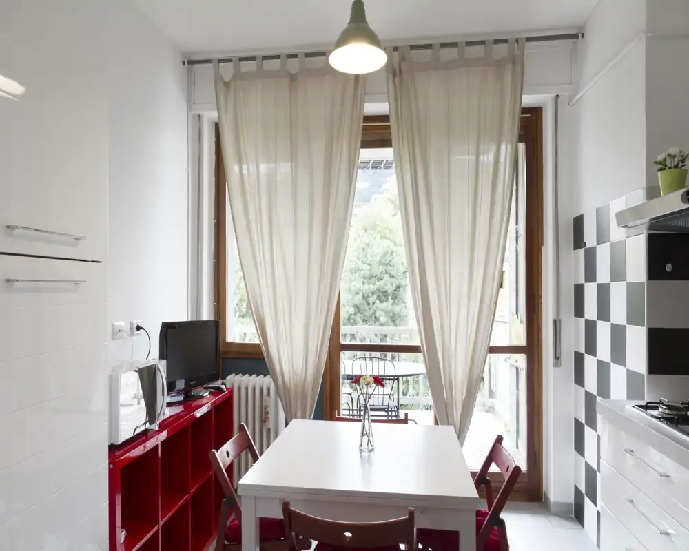 Cocina moderna y luminosa con mesa blanca central, sillas y estantería rojas, y una gran ventana que da a un pequeño balcón. Salpicadero de azulejos y electrodomésticos compactos para una cocina-comedor práctica y con estilo.