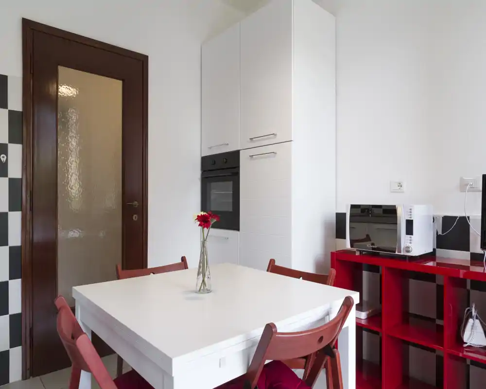 Rincón de cocina moderno y luminoso con mesa blanca, sillas y estantería rojas, horno empotrado y microondas — un espacio compacto y con estilo para comidas informales.
