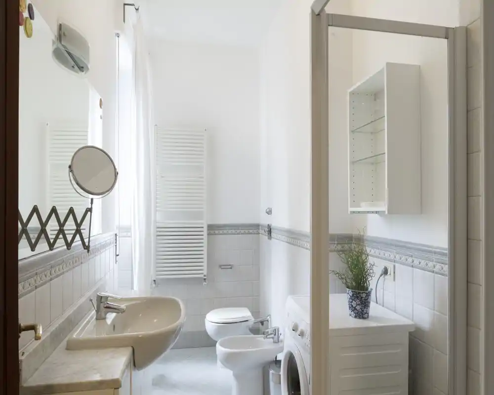 Baño luminoso y bien cuidado con lavabo, inodoro, bidé y cabina de ducha. Azulejos neutros y una pequeña lavadora crean un espacio funcional y limpio.