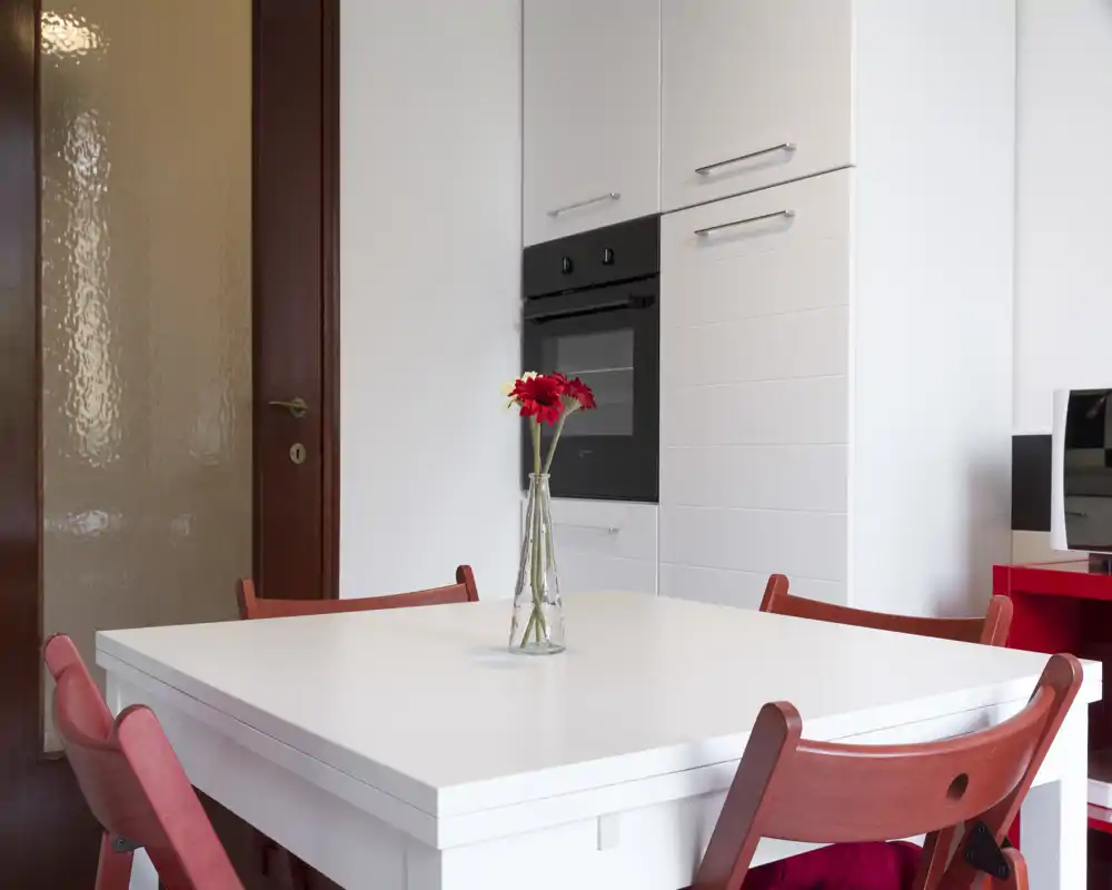 Rincón de comedor moderno y luminoso con mesa blanca, sillas rojas y un jarrón con flores: espacio limpio, minimalista y bien iluminado, ideal para comer.