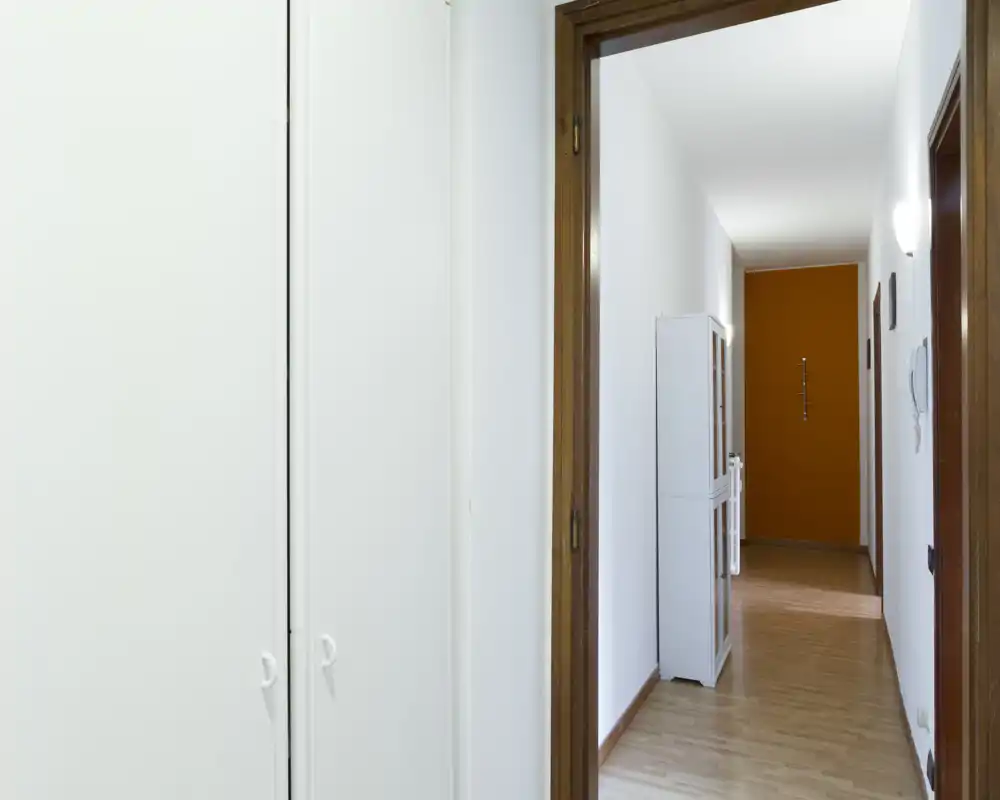 Vestíbulo de entrada luminoso y sencillo con suelo de madera, paredes blancas y un armario empotrado; se ve una pared naranja al fondo.