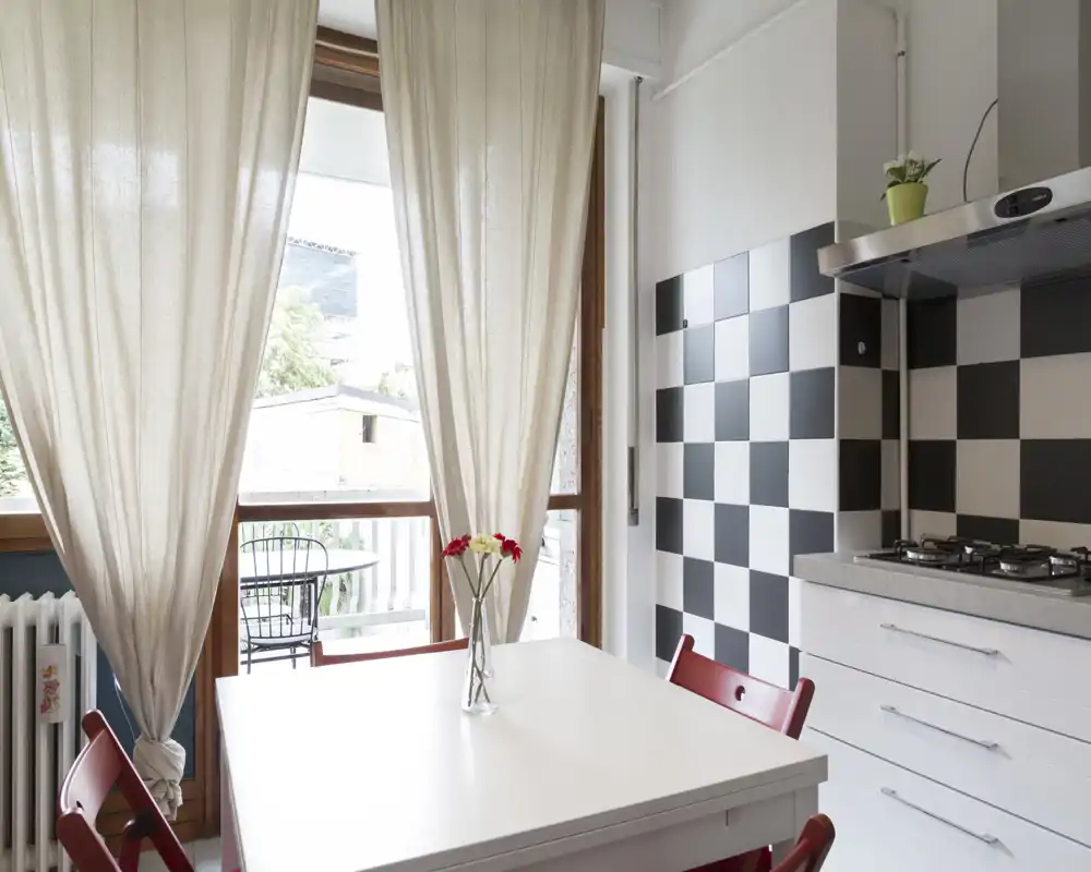 Esquina de cocina luminosa y moderna con mesa blanca, sillas rojas y salpicadero a cuadros; gran ventana/puerta que da a un pequeño balcón y aporta abundante luz natural.