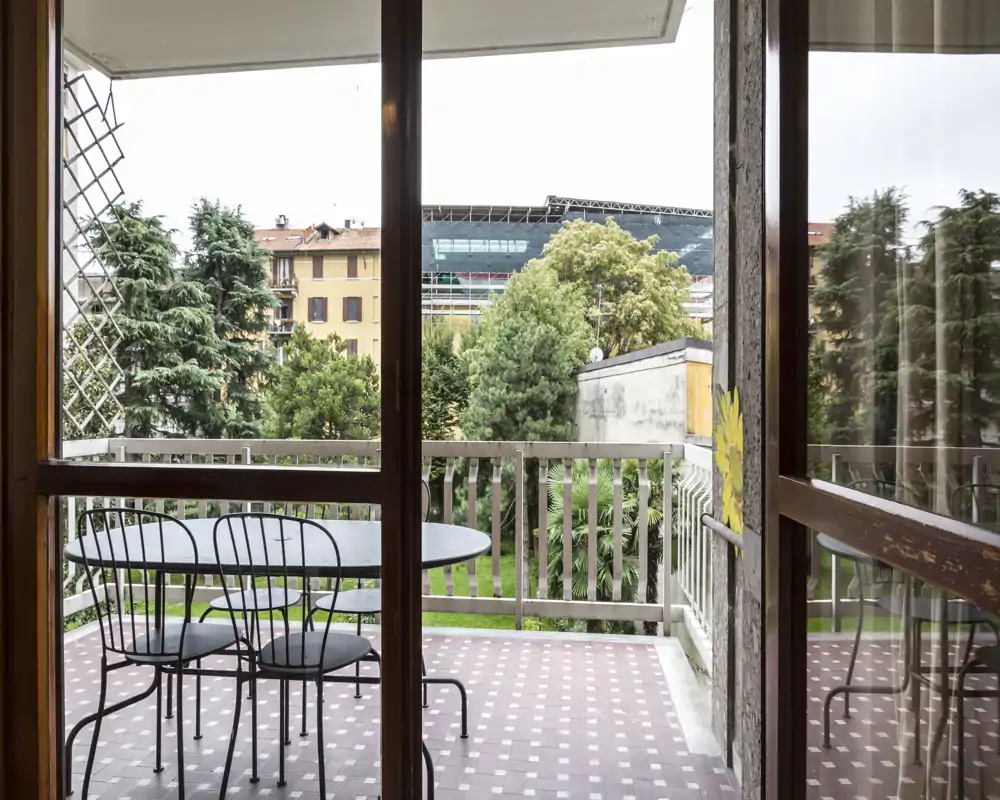 Balcón soleado con mesa redonda de metal y tres sillas a juego, suelo con baldosas y vistas a árboles y edificios cercanos — ideal para cenas al aire libre.