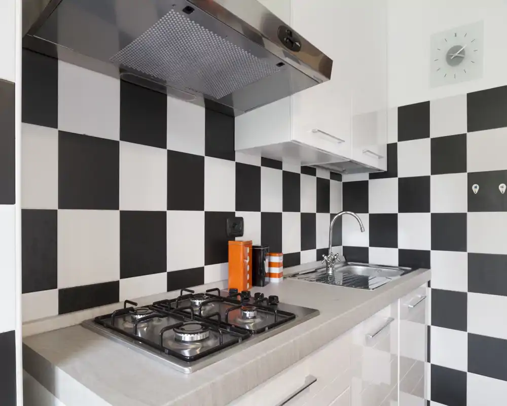 Cocina moderna y compacta con placa de gas, fregadero de acero inoxidable y azulejos en damero blanco y negro; encimera ordenada con pequeños botes decorativos.