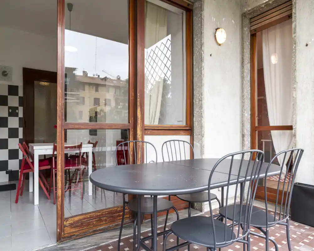 Balcón cubierto con mesa y sillas de metal negro, puertas de madera y cristal que comunican con el interior; ideal para comidas al aire libre y relajación.