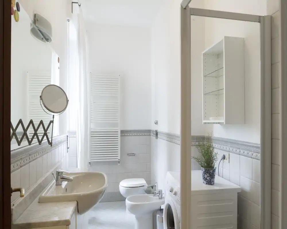 Baño luminoso y bien cuidado con lavabo, bidé, inodoro, plato de ducha y lavadora; azulejos neutros y radiador toallero alto que aportan sensación de limpieza y amplitud.