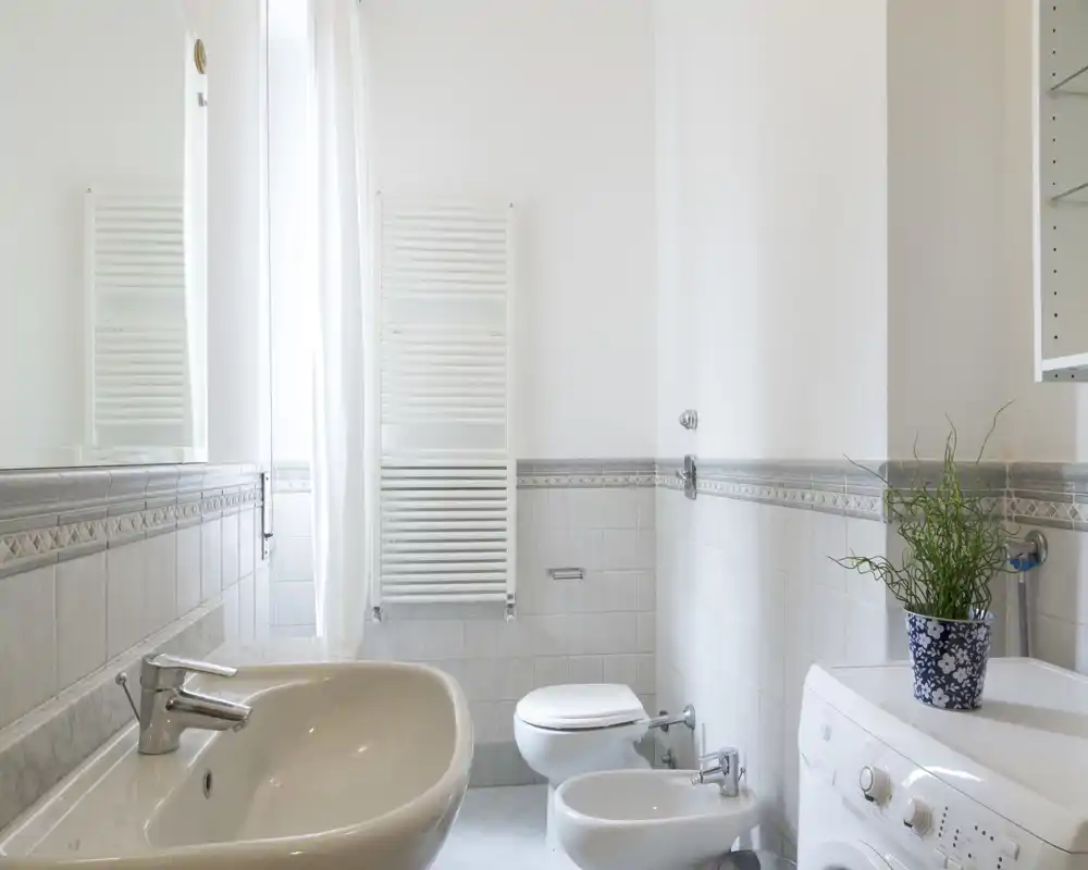 Baño luminoso y limpio con lavabo, bidé, inodoro y lavadora; azulejos claros y un radiador para toallas que crean una sensación fresca y aireada.