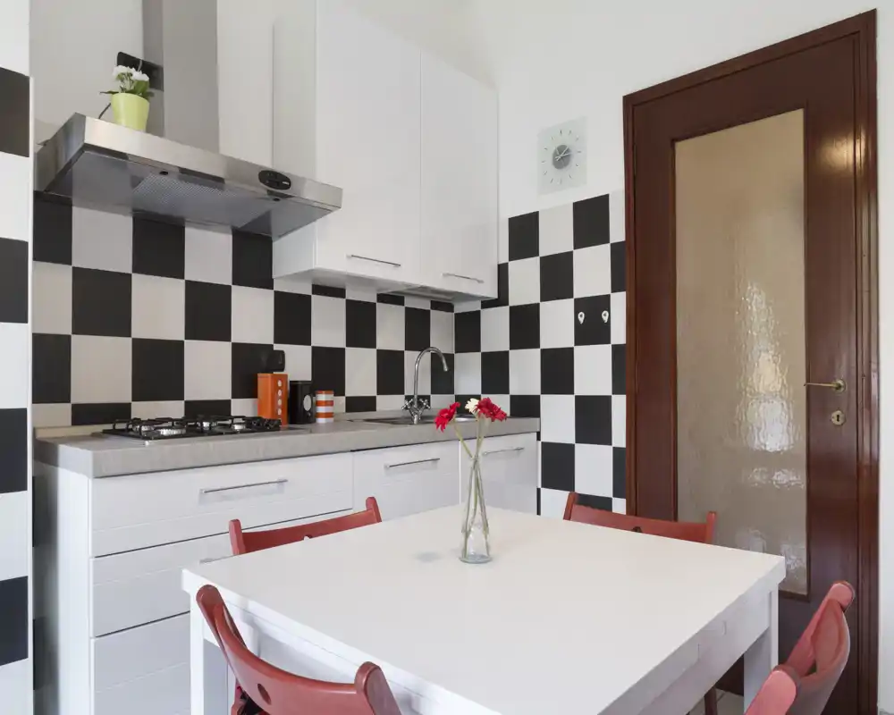 Cocina moderna y luminosa con mesa blanca y sillas rojas, salpicadero de cuadros, placa de gas y encimera con fregadero: un espacio compacto y con estilo para comidas informales.