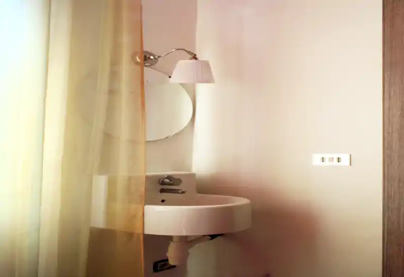 Compact bathroom corner with a wall-mounted round sink, small mirror and wall lamp behind a sheer curtain, softly lit and tidy.