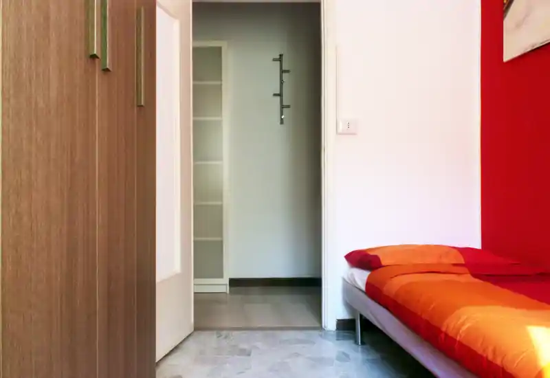 Compact bedroom with a single bed dressed in warm orange and red bedding, a built-in wardrobe on the left and a doorway leading to a small hall; bright, tidy and practical for a short stay.
