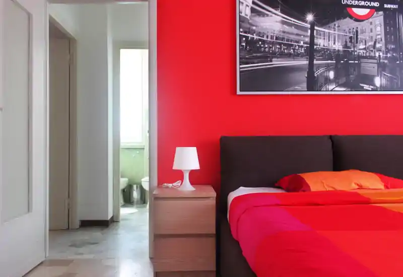 Bright, modern bedroom with a dark upholstered headboard and a vibrant red-and-orange duvet. A wooden bedside table with a lamp and a framed black-and-white photo complete the styled composition.
