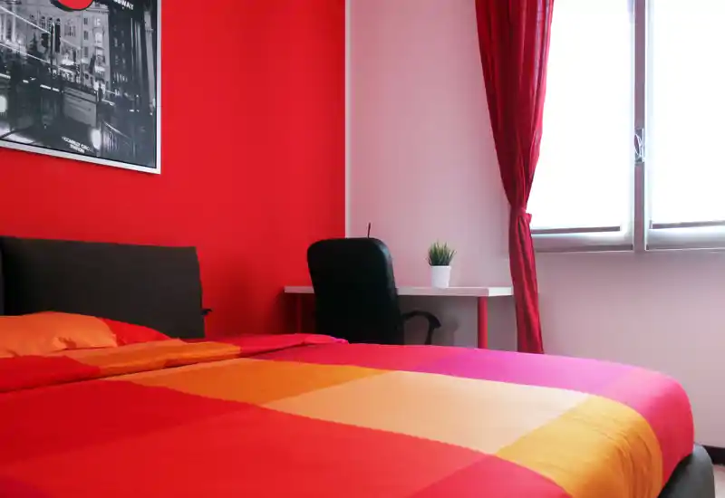 Bright, warm bedroom with a large double bed dressed in colorful bedding, a small desk and chair by the window and modern wall art.