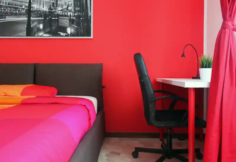 Bright, colorful bedroom with a large bed, bedside headboard and a compact workspace with chair and desk beside a window.