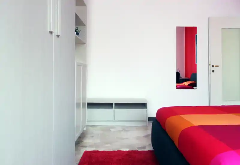 Bright, modern bedroom with a colorful red-orange bedspread, simple low furniture and a clean minimalist layout — ideal for a contemporary rental.