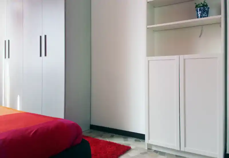 Bright, modern bedroom corner with a partially visible red bed, white wardrobe and shelving — clean lines and warm textiles create a simple, inviting space.