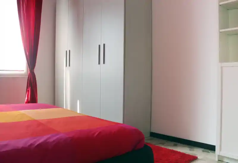 Bright, tidy bedroom with a large colorful bed in the foreground, a wardrobe along the wall and a window with deep-red curtains providing natural light.