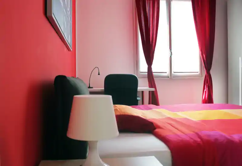 Bright, colorful bedroom with a double bed in the foreground, bedside lamp and a desk by the window with red curtains letting in natural light.