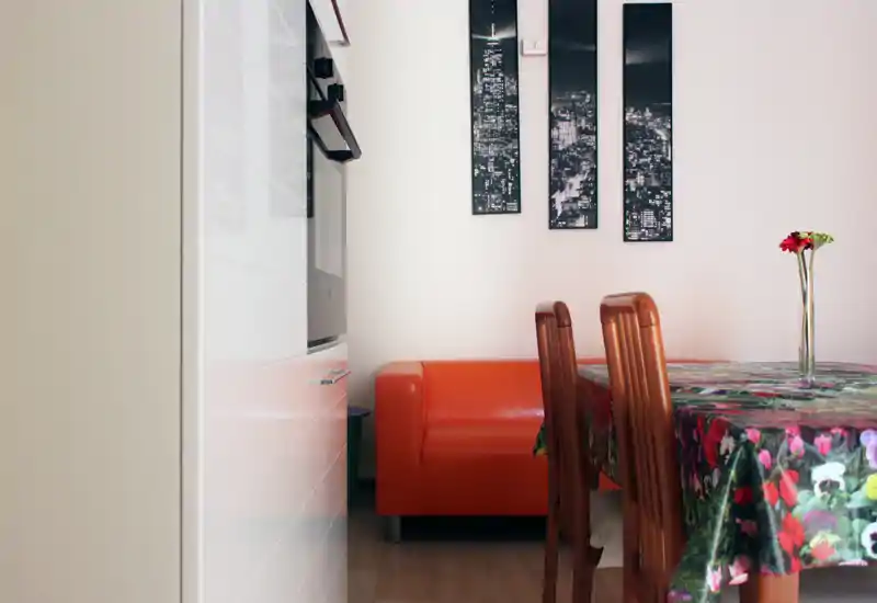 Compact living/dining corner with a bright orange sofa, wooden dining chairs and a floral tablecloth; modern wall art and a single vase create a welcoming, space-efficient layout.