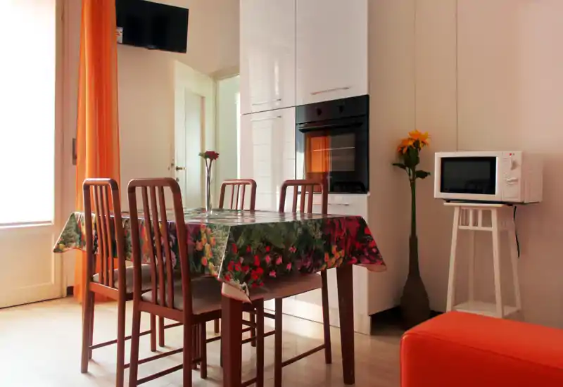 Bright, cozy dining area with a wooden table for four, floral tablecloth, and warm orange accents; built-in oven and microwave visible in the background.