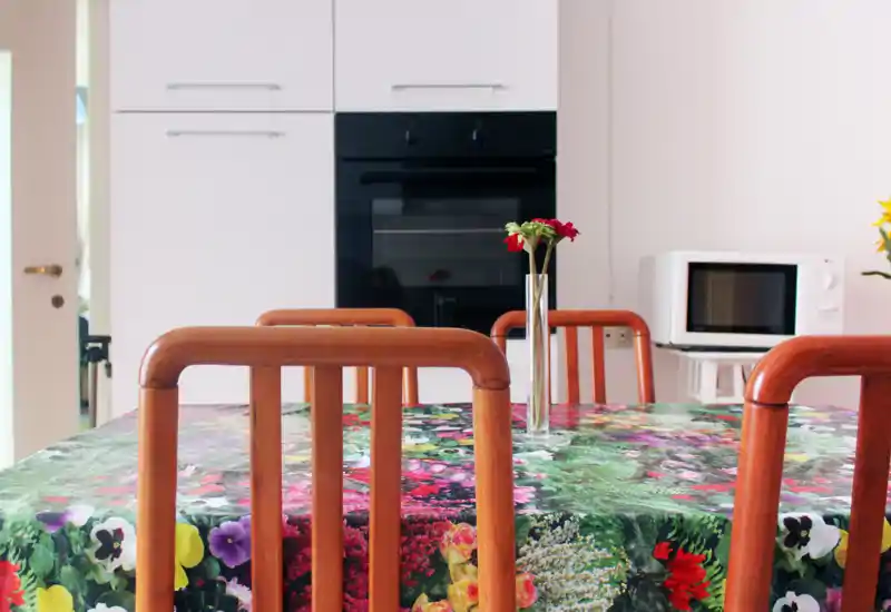 Zona pranzo luminosa con tovaglia floreale, sedie in legno e un piccolo vaso al centro; in background si vedono elettrodomestici della cucina (forno, microonde).