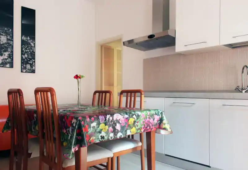 Bright, well-styled dining area with a floral tablecloth, wooden chairs and a compact kitchenette alongside — ideal for casual meals.