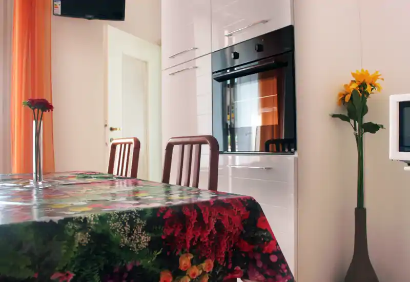 A bright, welcoming dining area with a floral tablecloth, two wooden chairs and decorative flowers, adjacent to built-in kitchen cabinets and oven.
