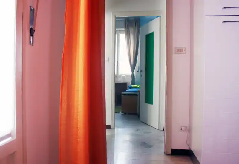 View through a doorway into a small bedroom with a visible bed in the distance, framed by an orange curtain and a bright window.