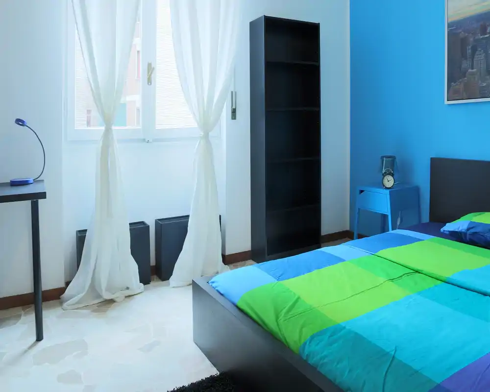 Camera da letto luminosa e moderna con un letto matrimoniale colorato, comodino con lampada e sveglia, libreria e ampia finestra con tende leggere che lasciano entrare luce naturale.