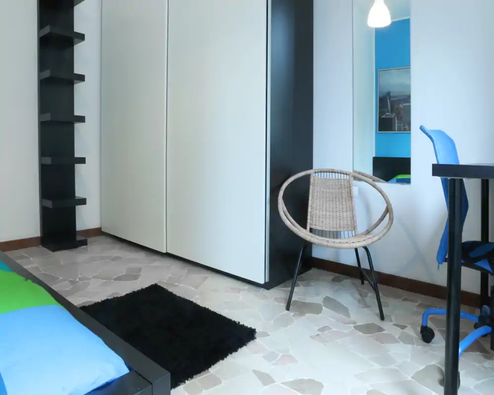 Angolo camera moderno e luminoso con bordo del letto visibile, grande armadio, sedia in rattan e una scrivania piccola — stile minimal e pulito, ideale per annunci.