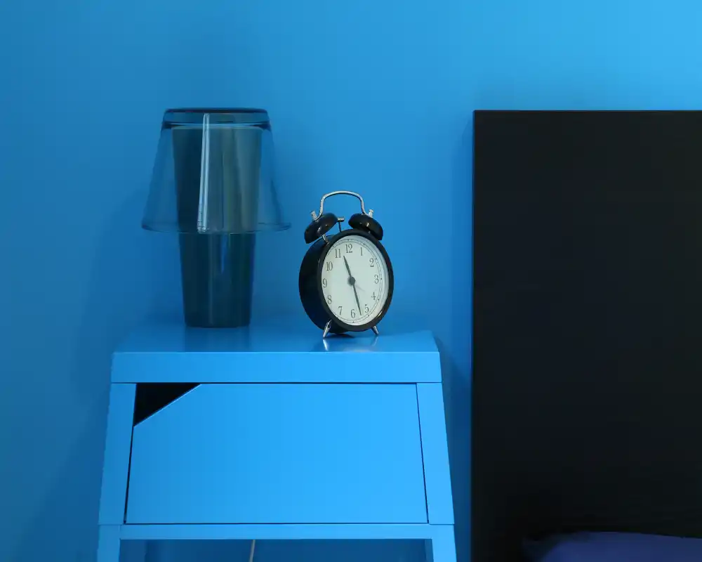 Dettaglio nitido del comodino in una camera blu: una lampada moderna e una sveglia classica su un comodino azzurro accanto a parte della testiera.