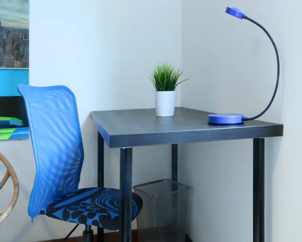 Angolo di lavoro compatto con una scrivania nera semplice, sedia ergonomica blu, piccola pianta in vaso e lampada da tavolo flessibile — luminoso, ordinato e adatto allo studio o al lavoro da remoto.