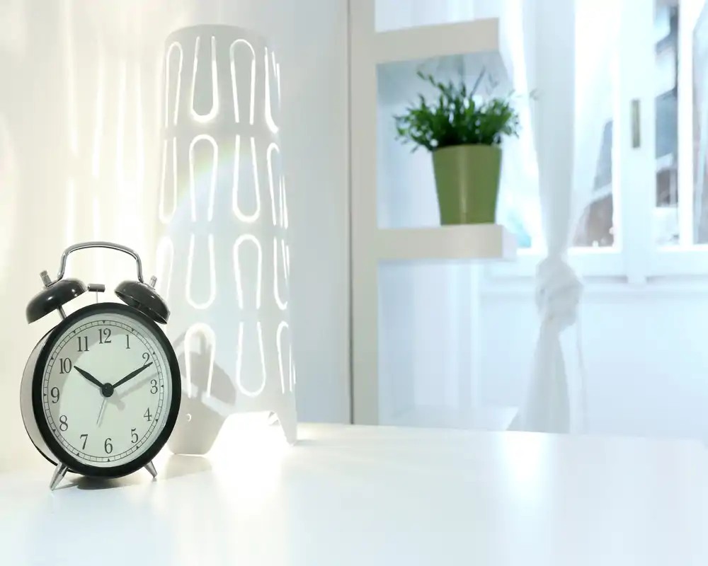 Escena de mesita de noche minimalista y luminosa con un despertador negro clásico, una lámpara blanca decorativa y una pequeña planta en estantería junto a una ventana soleada.