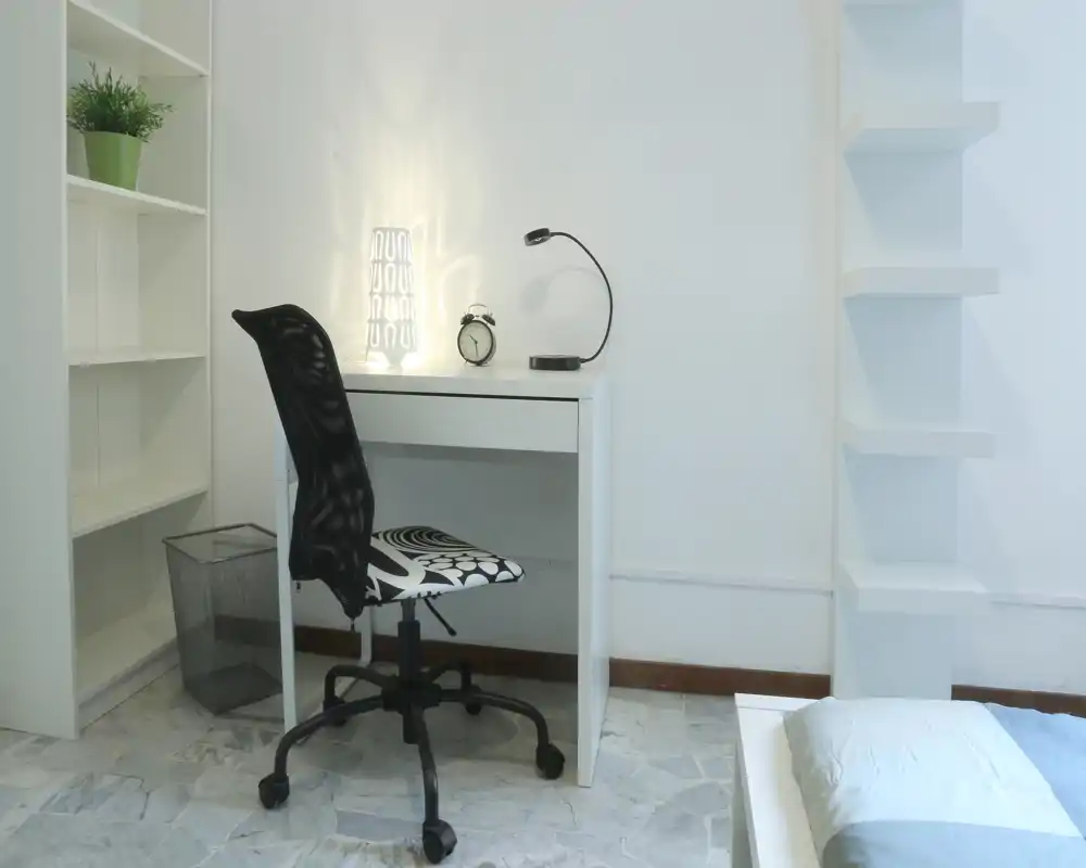 Rincón de dormitorio con un pequeño escritorio blanco, silla giratoria y parte de la cama visible. Decoración simple y limpia con lámpara y estanterías.
