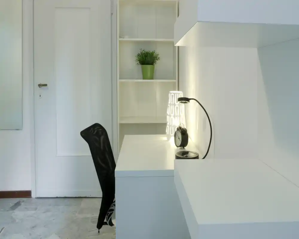 Rincón de trabajo compacto y moderno con escritorio blanco, lámpara de escritorio y silla negra; decoración mínima y luz suave crean un área limpia y funcional.