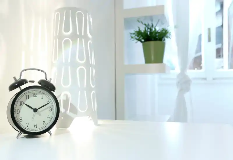 Escena de mesita de noche minimalista y luminosa con un despertador negro clásico, una lámpara blanca decorativa y una pequeña planta en estantería junto a una ventana soleada.