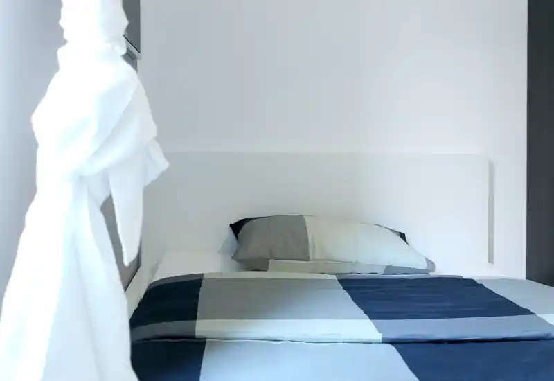 Dormitorio luminoso y minimalista con una cama bien hecha en tonos gris y azul marino; cabecero sencillo y luz natural que crean un espacio tranquilo.