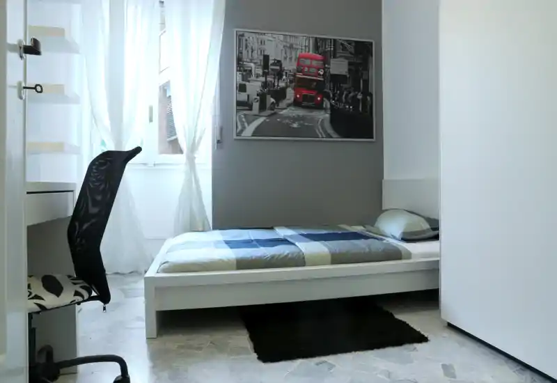 Dormitorio individual luminoso y minimalista con cama baja blanca, colcha a cuadros y luz natural desde la ventana; un escritorio con silla crea un pequeño espacio de trabajo.