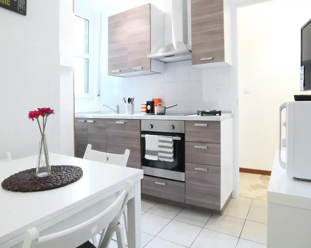 Cocina moderna y luminosa con muebles efecto madera, horno empotrado, placa de gas y una pequeña mesa — espacio limpio y bien iluminado, ideal para alquileres de corta estancia.