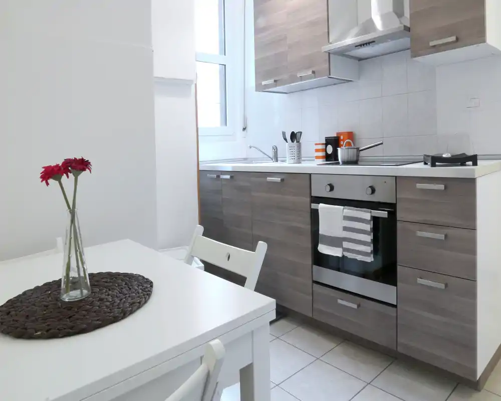 Cocina compacta y moderna con muebles imitación madera, horno integrado y una pequeña mesa; luz natural y detalles decorativos sencillos que la hacen acogedora.