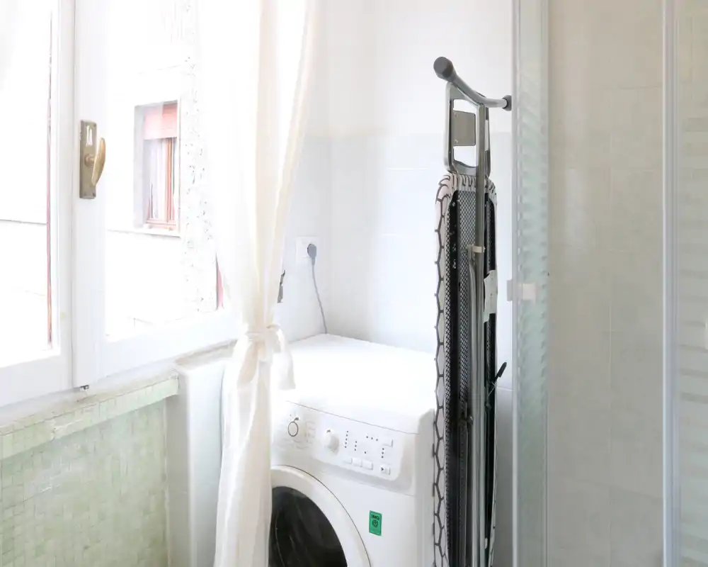 Rincón de baño con lavadora de carga frontal junto a una ducha y una ventana luminosa; área práctica de baño/lavandería.