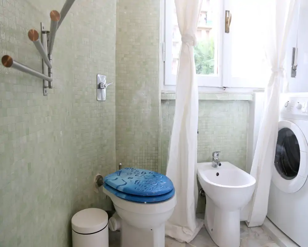 Baño compacto con inodoro, bidé y lavadora junto a una ventana luminosa; azulejos mosaico verde claro y cortinas claras que dan una sensación fresca y aireada.